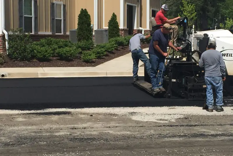 Paving crew running a paver alongside a commercial building, laying a fresh mat of hot-mix asphalt