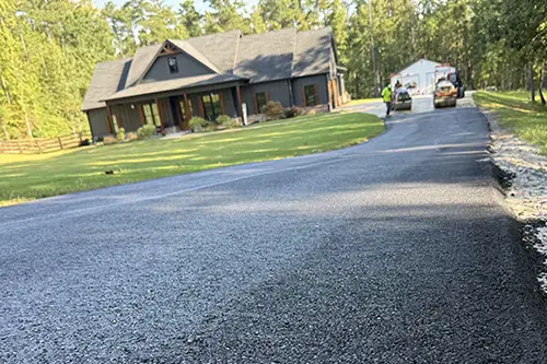 Finished asphalt driveway curving up to a house, with a roller and crew still working at the top end