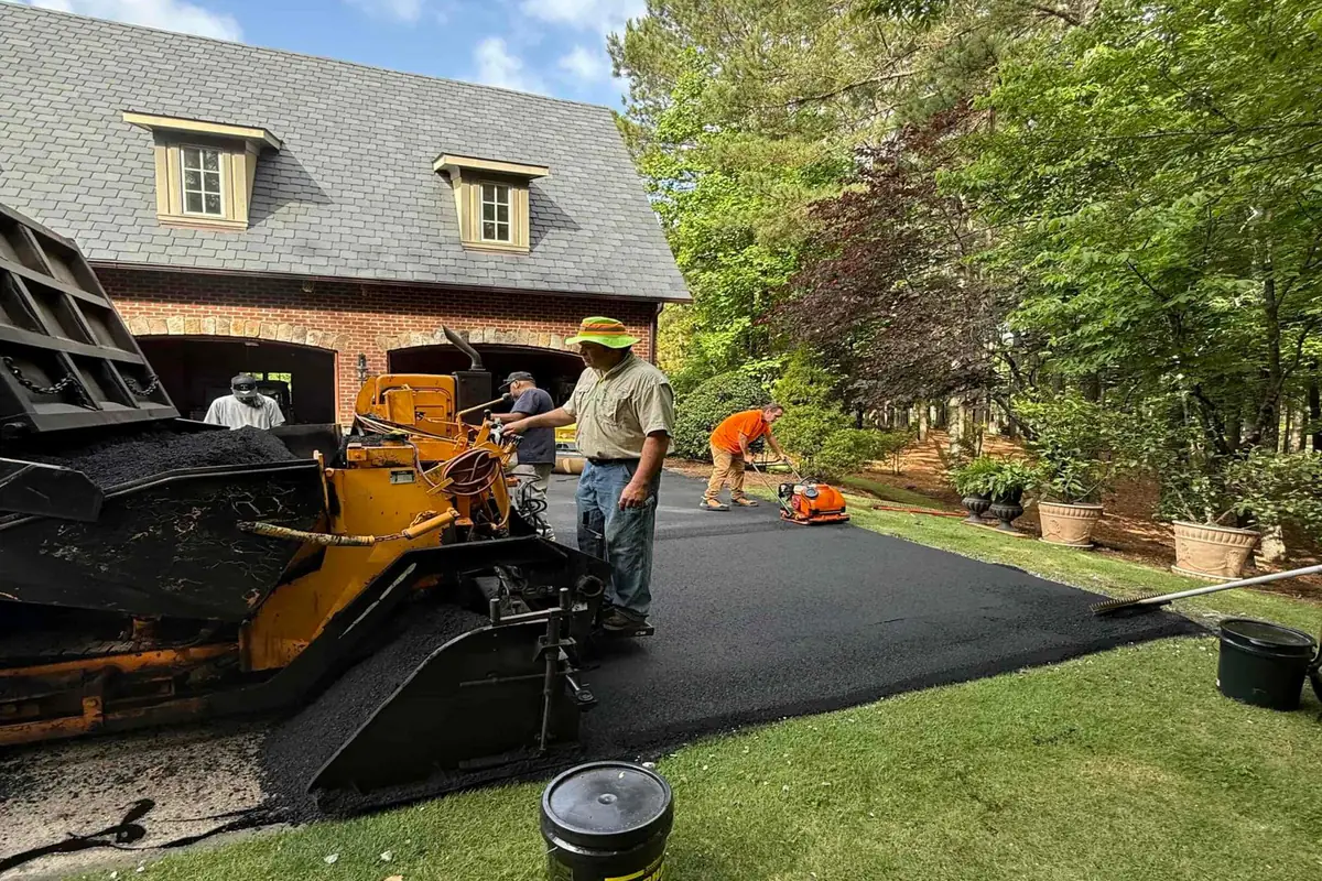 Paver laying a mat of hot-mix asphalt on a residential driveway in front of a brick house, with a crew member working the edge by hand