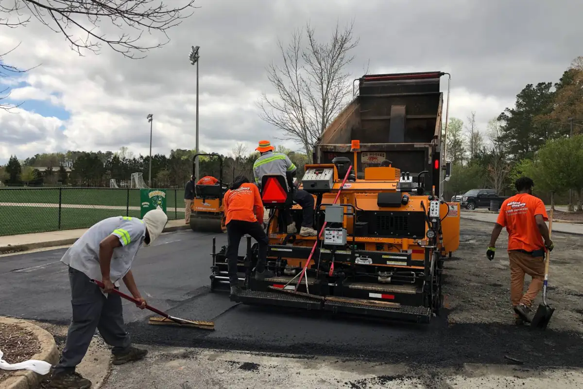 Paving crew placing hot-mix asphalt on a commercial lot, with a paver, a haul truck tipping into the hopper, and a worker raking the edge