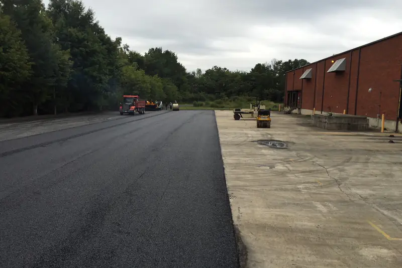 Newly paved truck court running alongside a brick warehouse, with paving equipment working at the far end