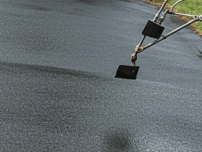 Spray wand applying black sealer across an asphalt driveway, with the wet leading edge visible against the dry surface ahead of it